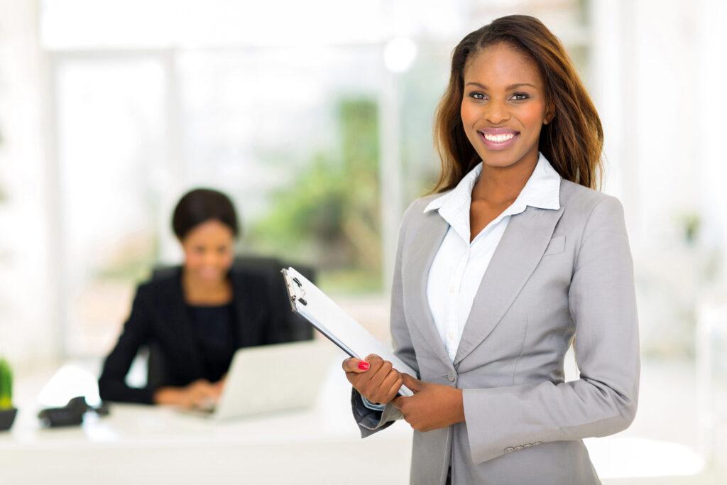 Rhode Island Inventors: Get Your $5,000 1 young woman in business suit holding papers, smiling, with another woman in background, working at a desk